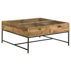Square coffee table with honey brown wood top, glass inset, two drawers, and black metal frame base.